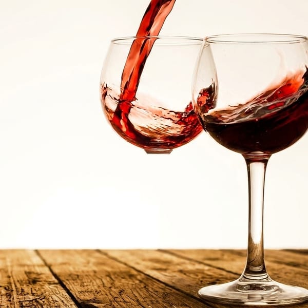 Red wine pouring into a glass, splashing, next to another full glass on a wooden table.