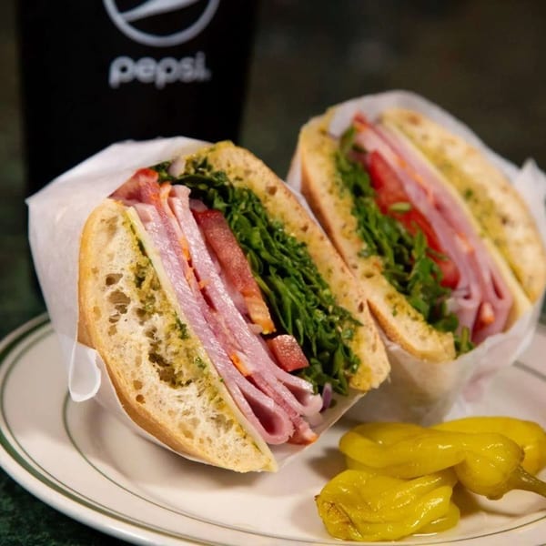 A deli sandwich with ham, cheese, lettuce, and tomato, served with peppers and a Pepsi.