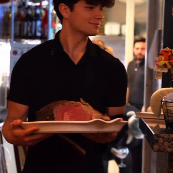Smiling server in a black shirt and apron carrying a large platter with a roasted prime rib.
