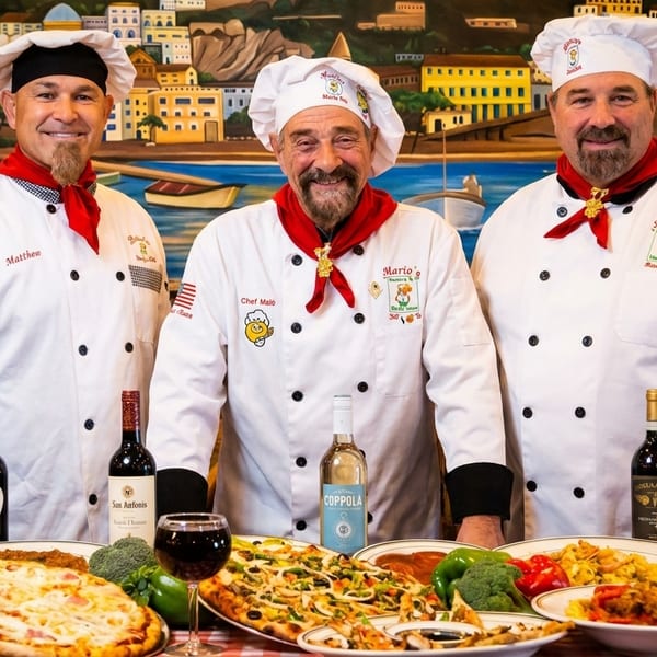 Three smiling chefs in white uniforms and red scarves stand behind a table of Italian food and wine.