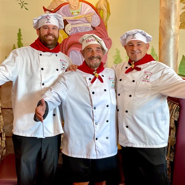 Three smiling chefs in white uniforms and red scarves stand in a restaurant with painted murals.