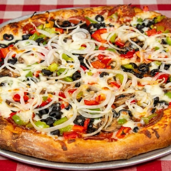 A whole supreme pizza with pepperoni, olives, peppers, onions, and mushrooms on a red checkered tablecloth.