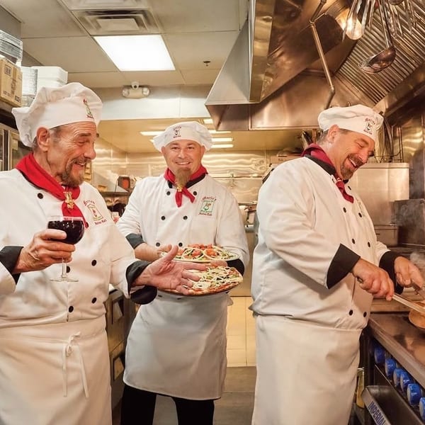Three smiling chefs in a bustling kitchen, one holding wine, another pizzas, and one cooking with flames.