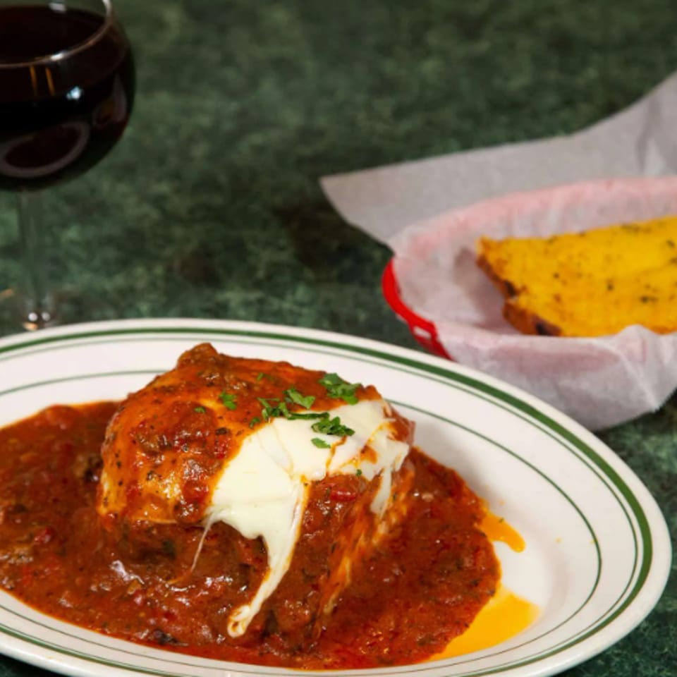 Cheesy enchilada covered in red sauce on a plate, with cornbread and a glass of red wine on a green tabletop.