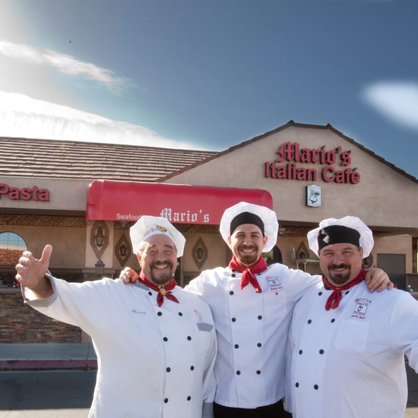 Three smiling chefs in white uniforms and red scarves stand proudly in front of Mario's Italian Cafe.
