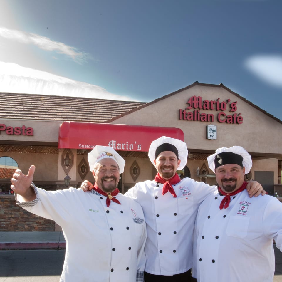 Three smiling chefs in white uniforms and red scarves stand proudly in front of Mario's Italian Cafe.