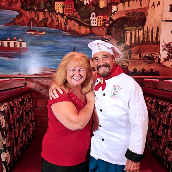A smiling chef and woman pose in a restaurant booth with a vibrant Italian mural background.