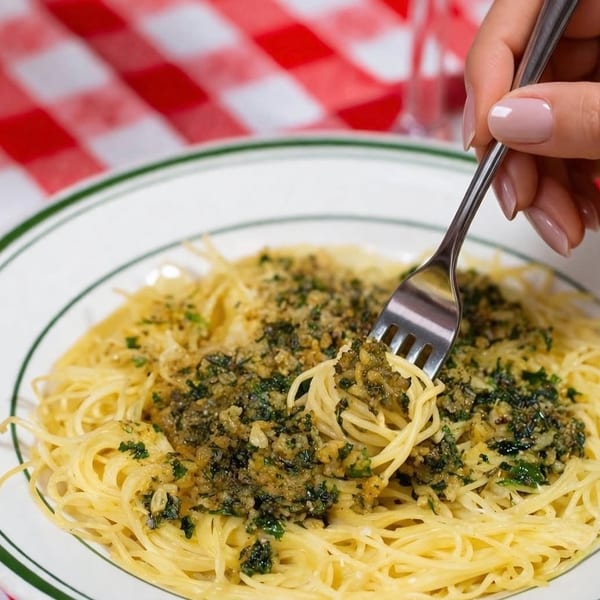 Plate of angel hair pasta topped with garlic, herbs, and breadcrumbs, while a hand twirls noodles with a fork.