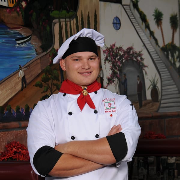 Smiling chef in white uniform and red scarf, arms crossed, in front of a painted mural.