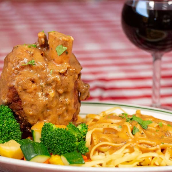 Plate of saucy meatloaf with broccoli, vegetables, and creamy pasta beside a glass of red wine.