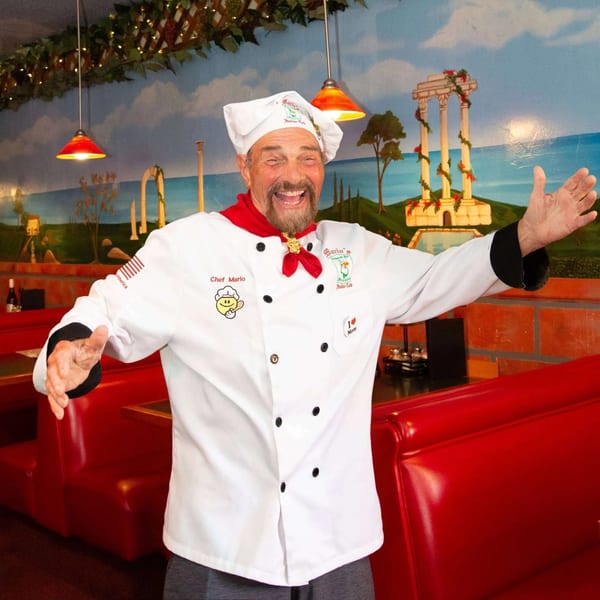 Chef Mario, a smiling man in a white chef's coat, stands in a restaurant with red booths.