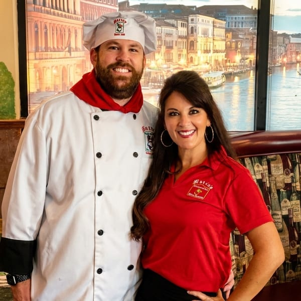 Smiling chef and woman in red shirt standing in a restaurant with a Venice canal mural.