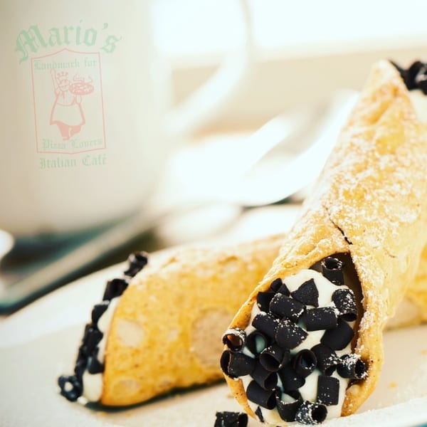 Cannoli filled with cream and topped with dark chocolate chips, displayed beside a branded Italian café cup.