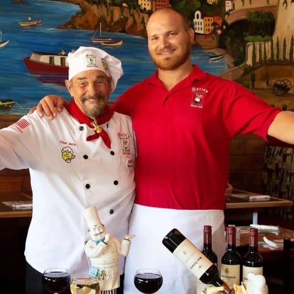 Chef Mario and a younger man in a red shirt stand in front of a vibrant Italian mural.