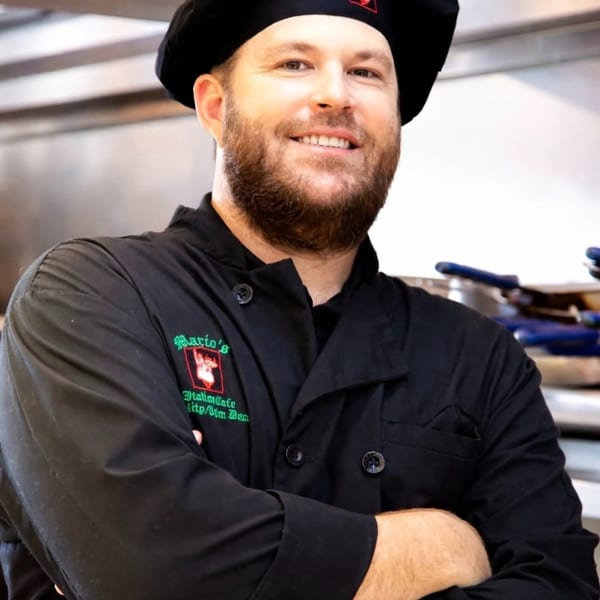Smiling chef in a black uniform and hat with the Mario's Italian Cafe logo, arms crossed in a kitchen.