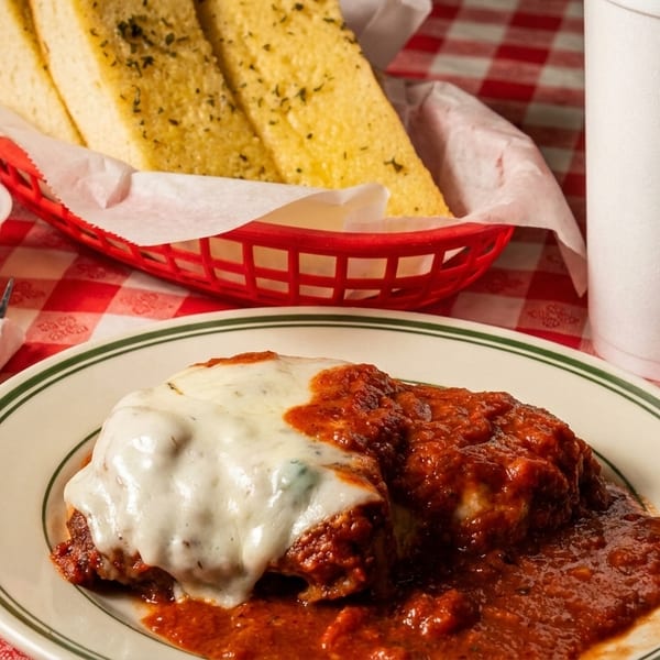 Chicken parmesan with melted cheese and red sauce, garlic bread, lemon wedges, and a drink on a red checkered tablecloth.