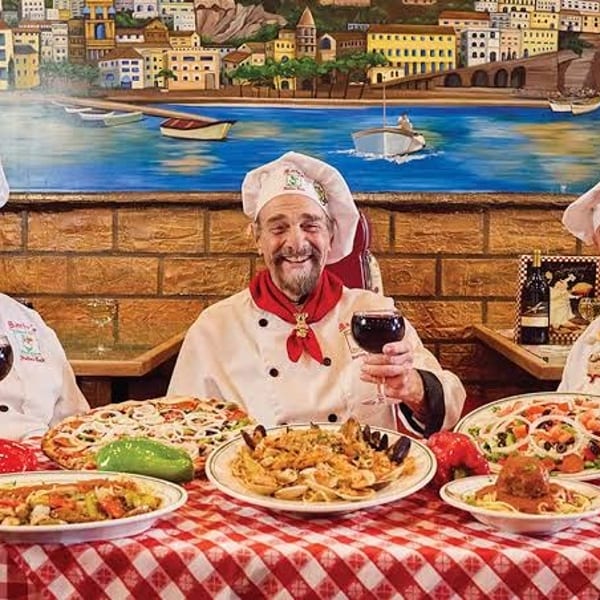 Three smiling chefs in white hats and red scarves toast with wine glasses at a table laden with Italian dishes.
