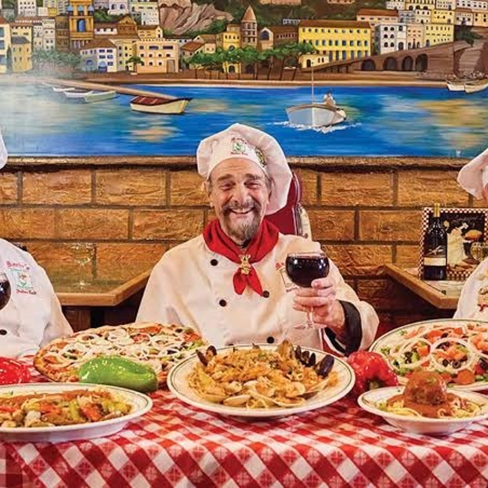 Three smiling chefs in white hats and red scarves toast with wine glasses at a table laden with Italian dishes.